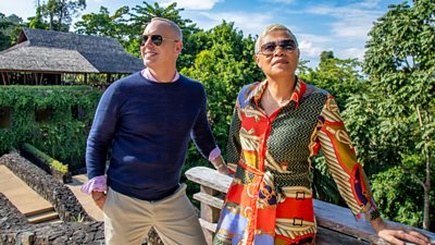 Presenters Rob Rinder and Monica Galetti stand in a carefully curated garden of a hotel. Dense, green woodland is visible in the background behind them. They both wear sunglasses and look off into the distance. 