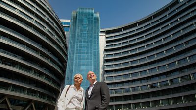 Presenters Rob Rinder and Monica Galetti stand outdoors at the base of a tall hotel in Shanghai. Monica looks to camera, while Rob looks up at the buildings in awe. 