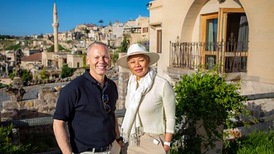 Presenters Rob Rinder and Monica Galetti smile to camera. Stood on a terrace, the skyline behind them shows stone buildings and structures across a hillside. 