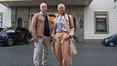 Presenters Rob Rinder and Monica Galetti stand on a stone plaza in front of a large hotel entrance. 