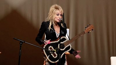 Dolly Parton sings into a microphone with a bedazzled guitar hung from a strap around her neck