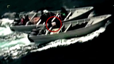 Two figures carry a white bag circled in red between two motorboats in the ocean from the point of view of a camera's scope.