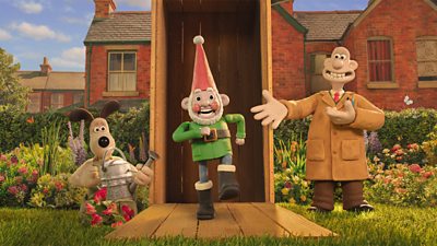 Scene from Wallace and Gromit: Vengeance Most Fowl. Wallace and Gromit stand either side of a large, open crate in a garden. Norbot the Smart Gnome walks out of the crate as Wallace gestures to the new invention. Gromit holds a watering can and looks at Norbot.