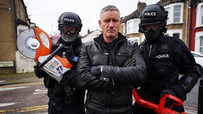 Billy Bingham with two police officers either side of him holding objects to break into a building