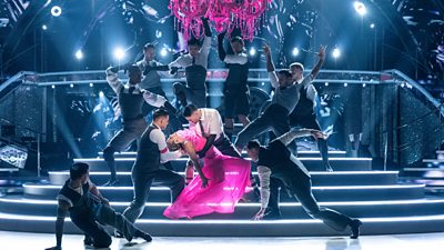 The Strictly ballroom is dramatically lit, with bright, white lights cutting through the darkness. Ten dancers dressed in white shirts and dark blazers and trousers strike varying poses on the steps of the dancefloor. In the centre of the image, a woman dressed in a vivid pink dress leans back in the arms of one of the dancers. 