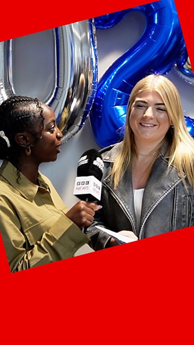 A woman interviews another woman as they stand in front of silver and blue balloons