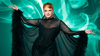 Drag Queen Chanel O'Conor wearing a black corset and black feathered neglige with her arms outstretched looking to the camera. Green smoke effect surrounding her