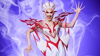 Drag Queen Kyran Thrax with one arm down and one arm up. She's wearing a pink and red outfit with big red jewelled spikes coming out of the hips, breast and shoulders. Her hair is white and red matching the outfit. Her mouth is open. Purple smoke in the background with a partial image of her in the smoke