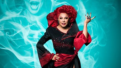 Drag Queen La Voix wearing a black dress with red jewels on it and clustered at the sleeve and across the breast and round the big pointed collar. Red sash across the waist that goes up into a sleeve. Her hand is up with one hand on her hip and red hair and eyebrows. Teals smoke effect in background with a partial image of her in it 