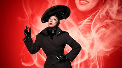 Drag Queen Saki Yew wearing a black coat with black leather gloves and leather collar/headpiece. She's got a black hat and nails coming out of her gloves. She's got one hand on her hip and one hand slightly pointing up. Red smoke effect in background with partial image of Saki in the smoke