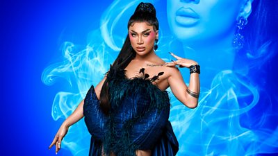 Drag Queen Zahirah Zapanta wearing a peacock feather bodice with material draping down the back of the garment. One hand is lightly touching her shoulder and the other is pointing down. Blue smoke effect iin the background with a partial image of Zahirah in the smoke