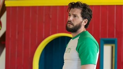 Henry (Kit Harington) in a white t shirt with green sleeves, stood in front of a brightly coloured wall. He looks to something offscreen in dismay. 