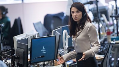 Yasmin (Marisa Abela) stands in front of a desk of computer screens wearing a headset. She has her arms raised to her sides in disbelief as she looks at something offscreen. 