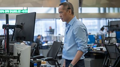 Eric (Ken Leung) stands in front of a computer screen wearing a telephone headset. He stares at the screen with a sombre expression. 