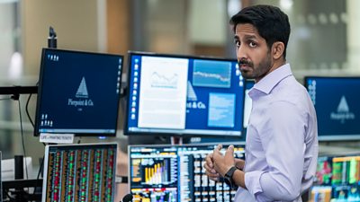 Rishi Ramdani (Sagar Radia) stands in front of several screens showing charts, documents and numerical data. He looks over his shoulder with a solemn expression at something offscreen. 