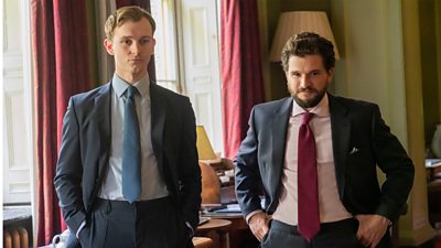 Robert (Harry Lawtey) and Henry (Kit Harington) stand next to each other wearing suits in a grand building with high ceilings and panelled walls. 