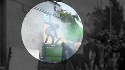 Man wearing a dark jacket and his hood up standing on top of a green wheelie bin that is billowing smoke.