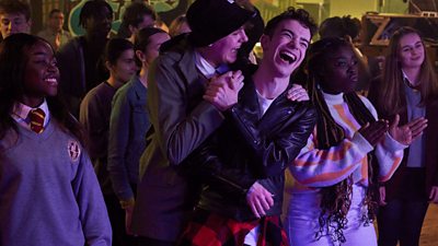 School teens in grey uniform with red and gold ties, two boys laughing and embracing, celebrating, while others are clapping in a darkened hall.