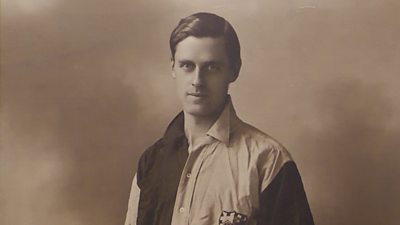 A black and white, historical photograph of a man in a multicoloured shirt standing against a plain background