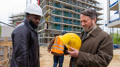 DI Russell Carter (Dipo Ola) and John ‘Ludwig’ Taylor (David Mitchell) at a building site in front of a large tower covered in scaffolding. DI Carter wears a hard-hat, while Ludwig holds a hard-hat and looks at it with a displeased expression. 