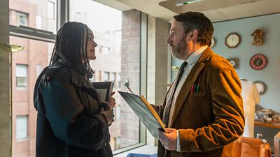 Izuka Hoyle as DS Alice Finch and David Mitchell as John ‘Ludwig’ Taylor/James Taylor talk to each other in a residential apartment while holding clipboards. The legs of a person lying on the floor can be seen in the background. 