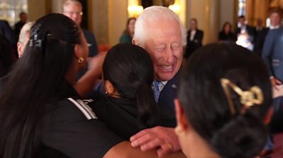 King Charles grins as three rugby players wearing black New Zealand t-shirts give him hug