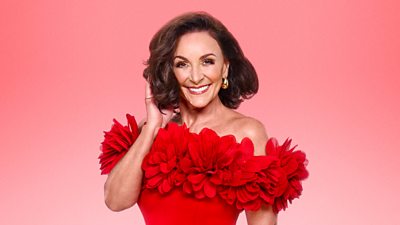 Shirley Ballas smiles for the camera wearing a bright red dress with a ruffled top. She holds one hand to the side of her hair.