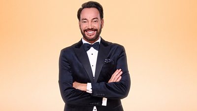 Craig Revel Horwood wears a dark tuxedo and bow tie and stands smiling for the camera with his arms folded