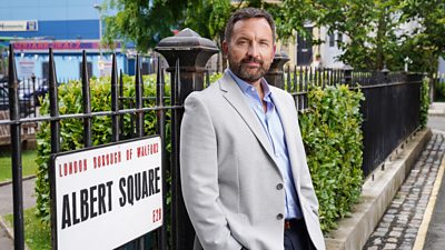 Jake Moon leans back against the railings in Albert Square, wearing a light grey suit jacket over a light blue shirt