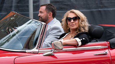 Jake Moon and Chrissie sit in a red convertible car. He looks forward while she leans her head to the side, looking towards the camera wearing dark sunglasses