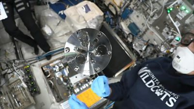 A circular metal part spins in zero gravity as two astronauts look on wearing masks and goggles.