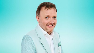 Chris McCausland is pictured side on wearing a light green suit and white shirt as he faces the camera