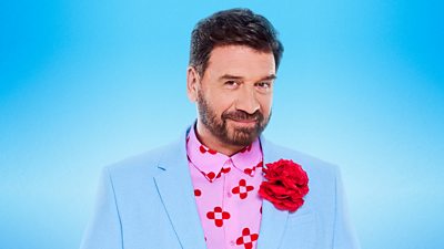 Nick Knowles wears a light blue suit jacket over a pink and red patterned shirt. There's a bright red flower on the lapel of his jacket.