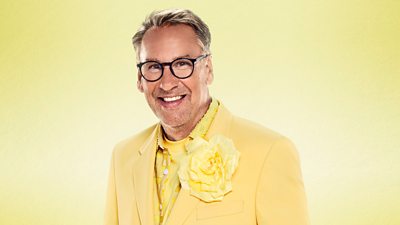 Paul Merson wears a bright yellow suit jacket and shirt, with a bright yellow flower on the lapel of the jacket. He smiles to camera wearing dark circular spectacles.