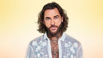 Pete Wicks looks straight down the lens of the camera, wearing a white translucent shirt, pattered with white flowers. His chest and neck tattoos are visible through the shirt.