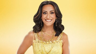 Dr Punam Krishan smiles for the camera wearing a bright gold/yellow dress, accessorised with gold necklaces and earrings.