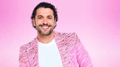 Shayne Ward wears a pink suit jacket dotted with gemstones in a stripy animal print style pattern over a white t-shirt and smiles to camera