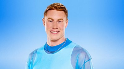 Tom Dean wears a white vest beneath a blue translucent sleeved top and sports a silver chain necklace, smiling to camera.