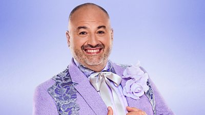 Wynne Evans wears a purple shirt with a ribbon tied in a bow at the collar, beneath a purple suit jacket with a sparkling embroidered pattern and a bright purple flower on the lapel.