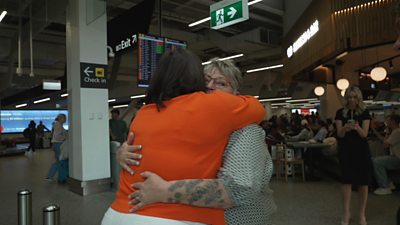 A woman wearing an orange top hugs another woman with tattoos inside an airport