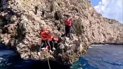 Several people stand on a rocky outcrop by the sea wearing life jackets. One holds a buoyancy ring