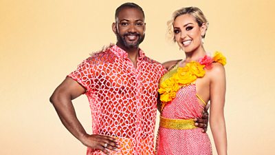 Jb and Amy stand smiling for the camera in red and yellow outfits. JB has an arm around Amy's waist.