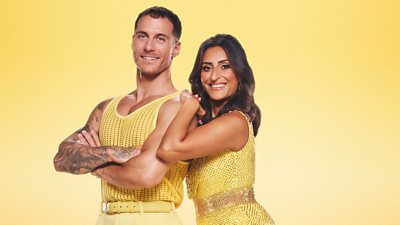 Punam and Gorka stand together in glittery yellow outfits. Gorka stands with his arms folded while Punam leans on his shoulder.