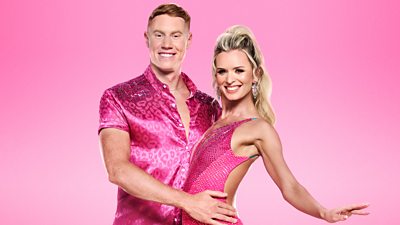 Tom and Nadiya pose in pink outfits. He holds her around the waist and they smile for the camera.