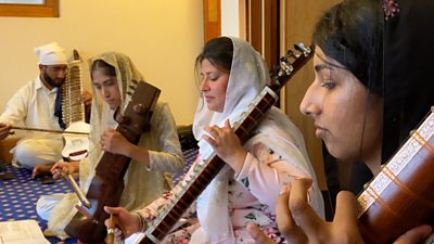 Three women have their heads covered with a scarf while playing a Dilruba which is an Indian string instrument.