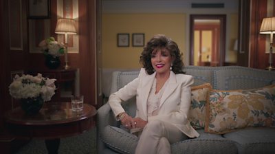 Dame Joan Collins in a white suit and blouse sitting on a sofa smiling 