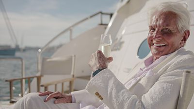 George Hamilton dressed in white on a yacht holding a glass of champagne and turning to camera and smiling 