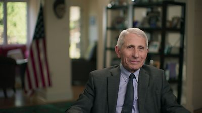 Dr. Anthony Fauci sitting down with a shirt and tie smiling. blurred living room in background with USA flag