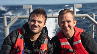 Scott Mills and Sam sit on a boat wearing life preservers, with the spray of water visible in the background