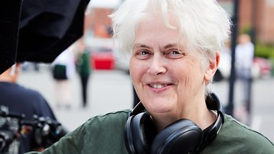 Sally Wainwright smiles from the set of Riot Women. She wears headphone around her neck. There are people with camera equipment in the background. 